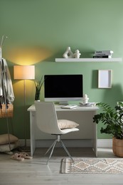 Photo of Home office. Comfortable workplace with desk, computer and lamp near light green wall in room