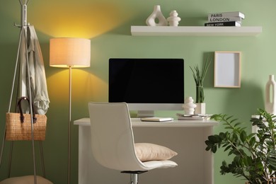 Photo of Home office. Comfortable workplace with desk, computer and lamp near light green wall in room