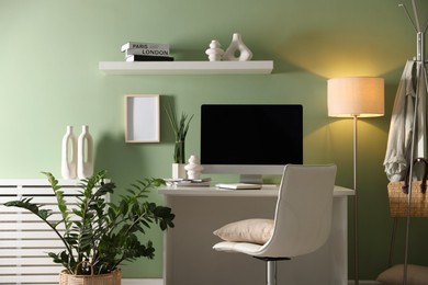 Photo of Home office. Comfortable workplace with desk, computer and lamp near light green wall in room