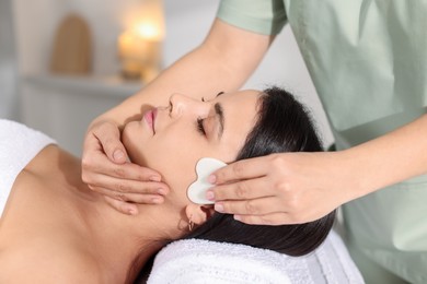 Photo of Young woman receiving facial massage with gua sha tool in spa salon