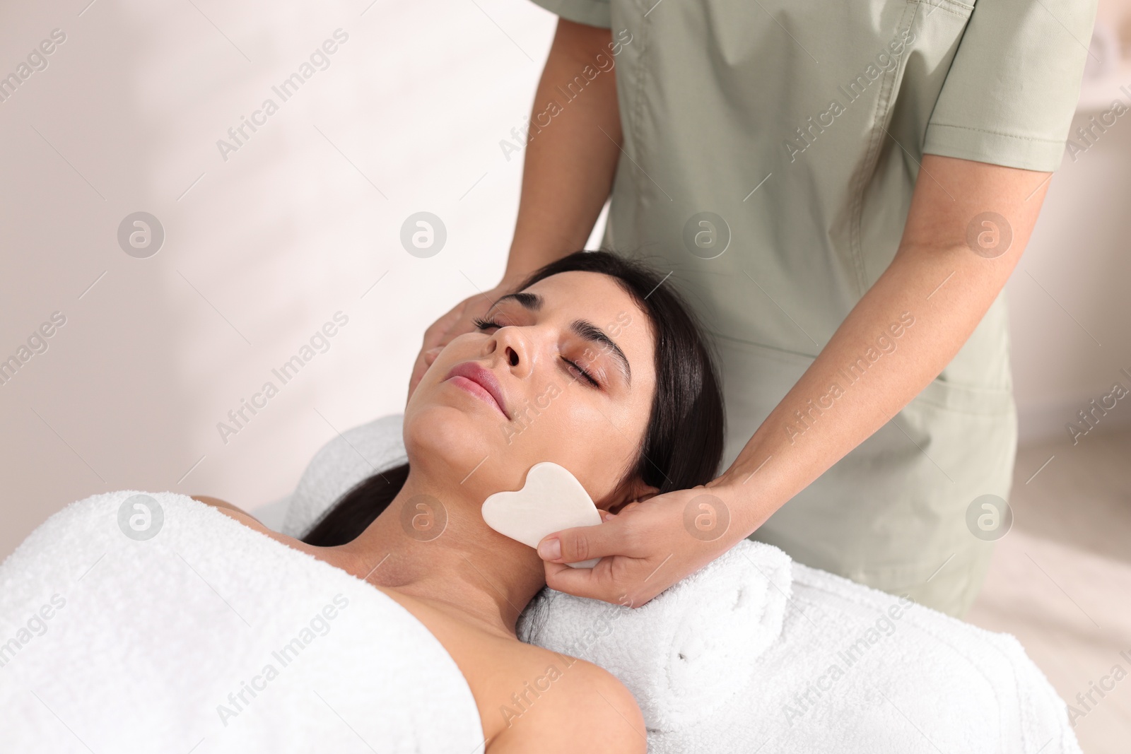 Photo of Young woman receiving facial massage with gua sha tool in spa salon