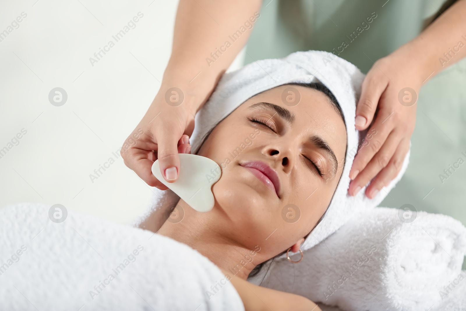 Photo of Young woman receiving facial massage with gua sha tool in spa salon