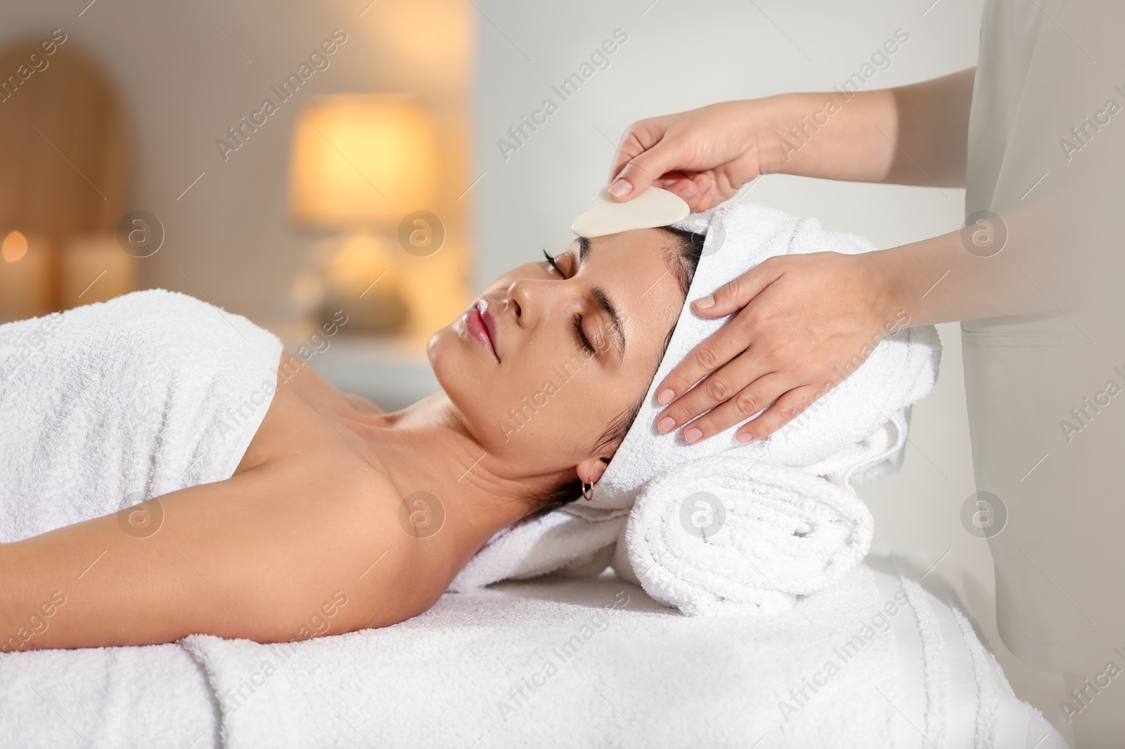 Photo of Young woman receiving facial massage with gua sha tool in spa salon