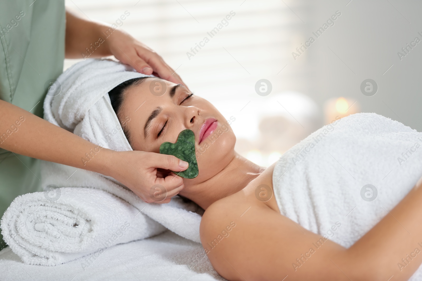 Photo of Young woman receiving facial massage with gua sha tool in spa salon