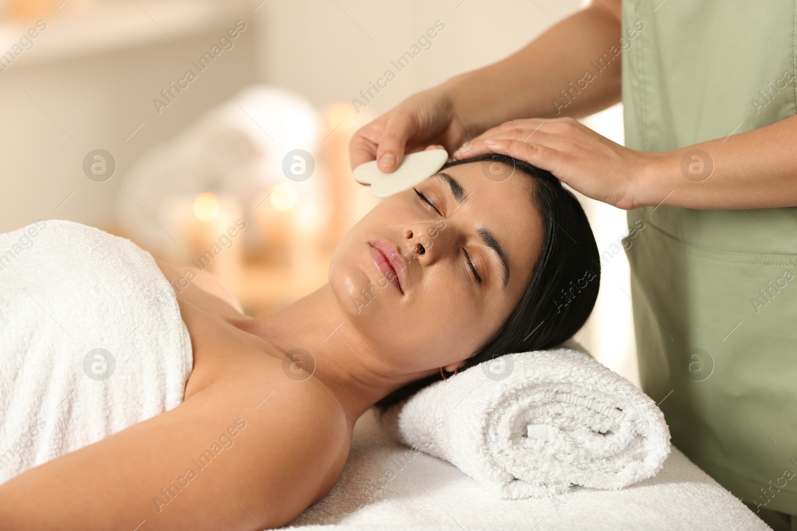 Photo of Young woman receiving facial massage with gua sha tool in spa salon