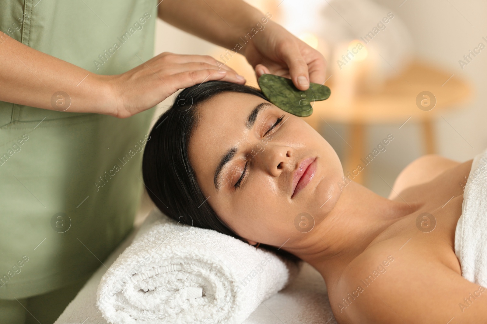 Photo of Young woman receiving facial massage with gua sha tool in spa salon