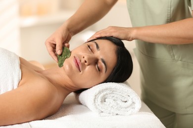 Photo of Young woman receiving facial massage with gua sha tool in spa salon