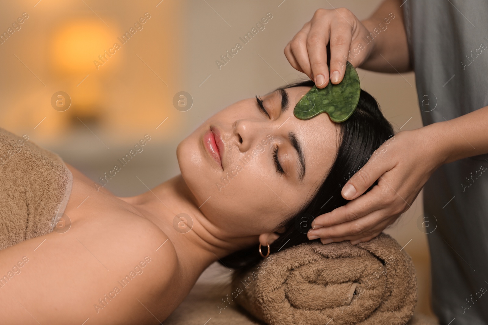 Photo of Young woman receiving facial massage with gua sha tool in spa salon