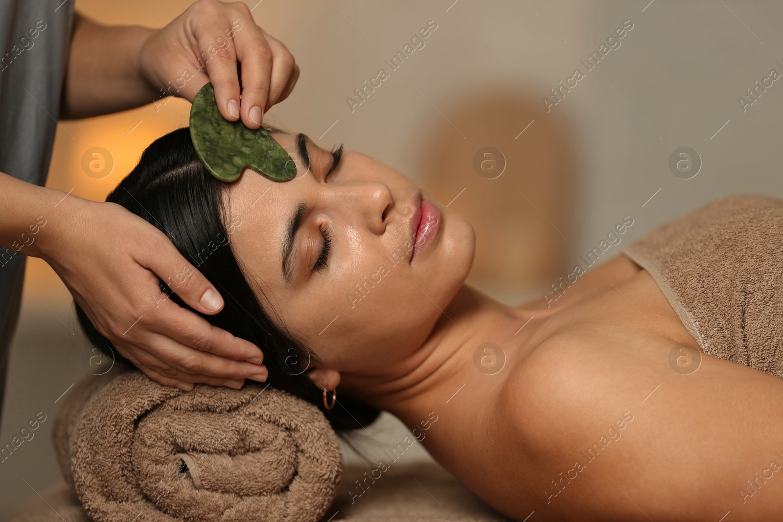 Photo of Young woman receiving facial massage with gua sha tool in spa salon