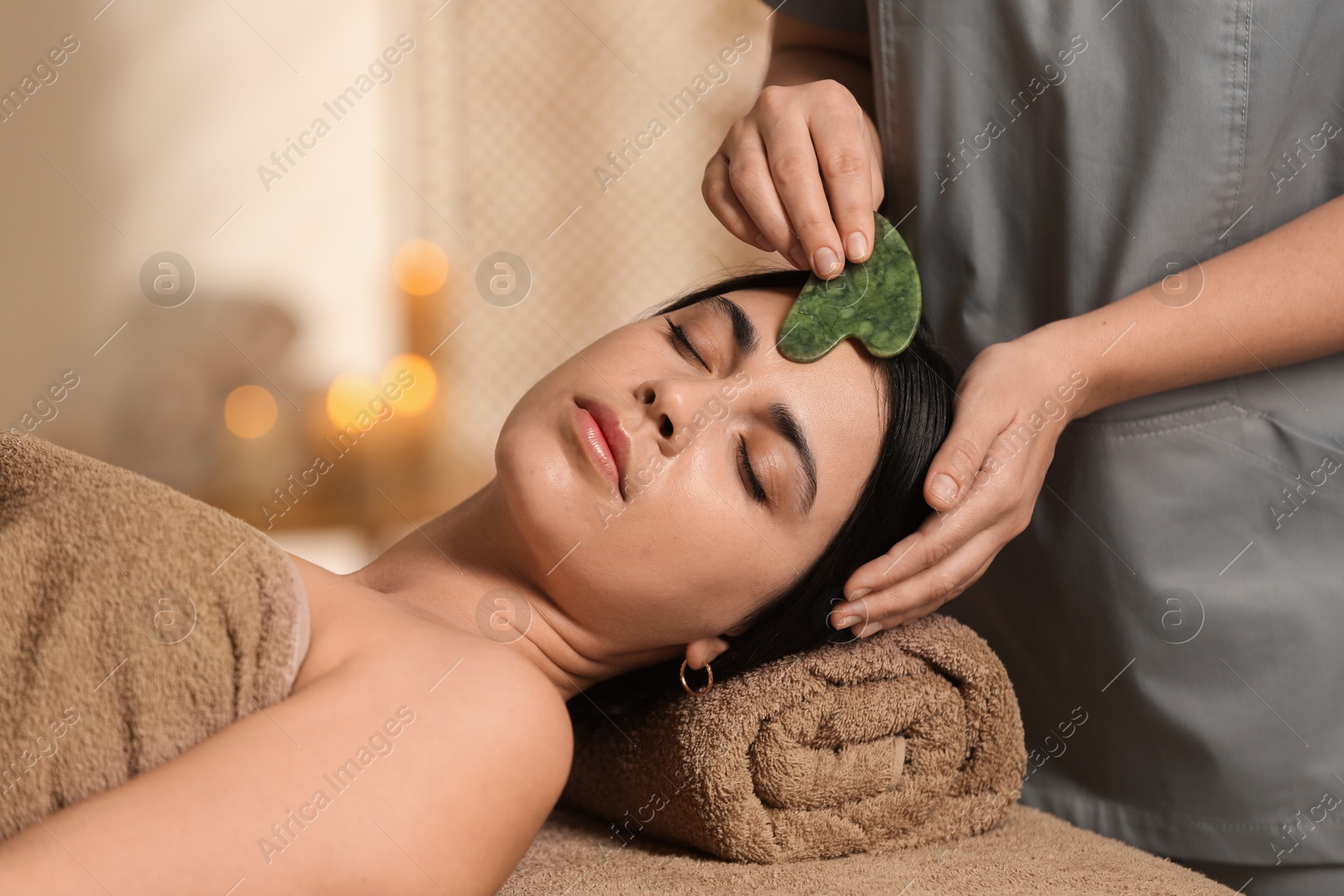 Photo of Young woman receiving facial massage with gua sha tool in spa salon