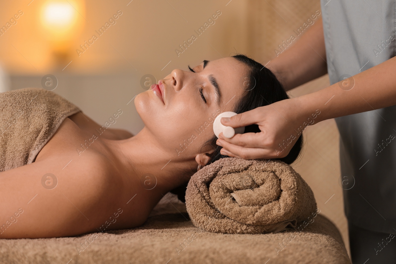 Photo of Young woman receiving facial massage with spa stone in salon