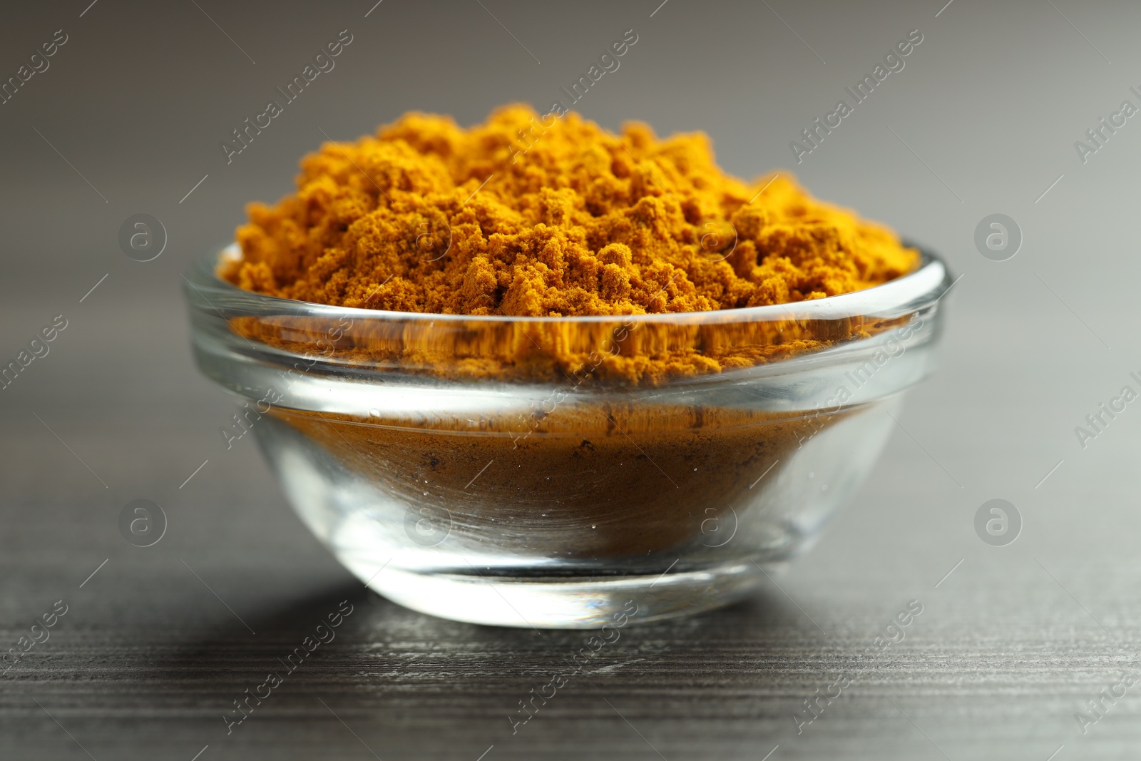 Photo of Turmeric powder in glass bowl on grey wooden table, closeup