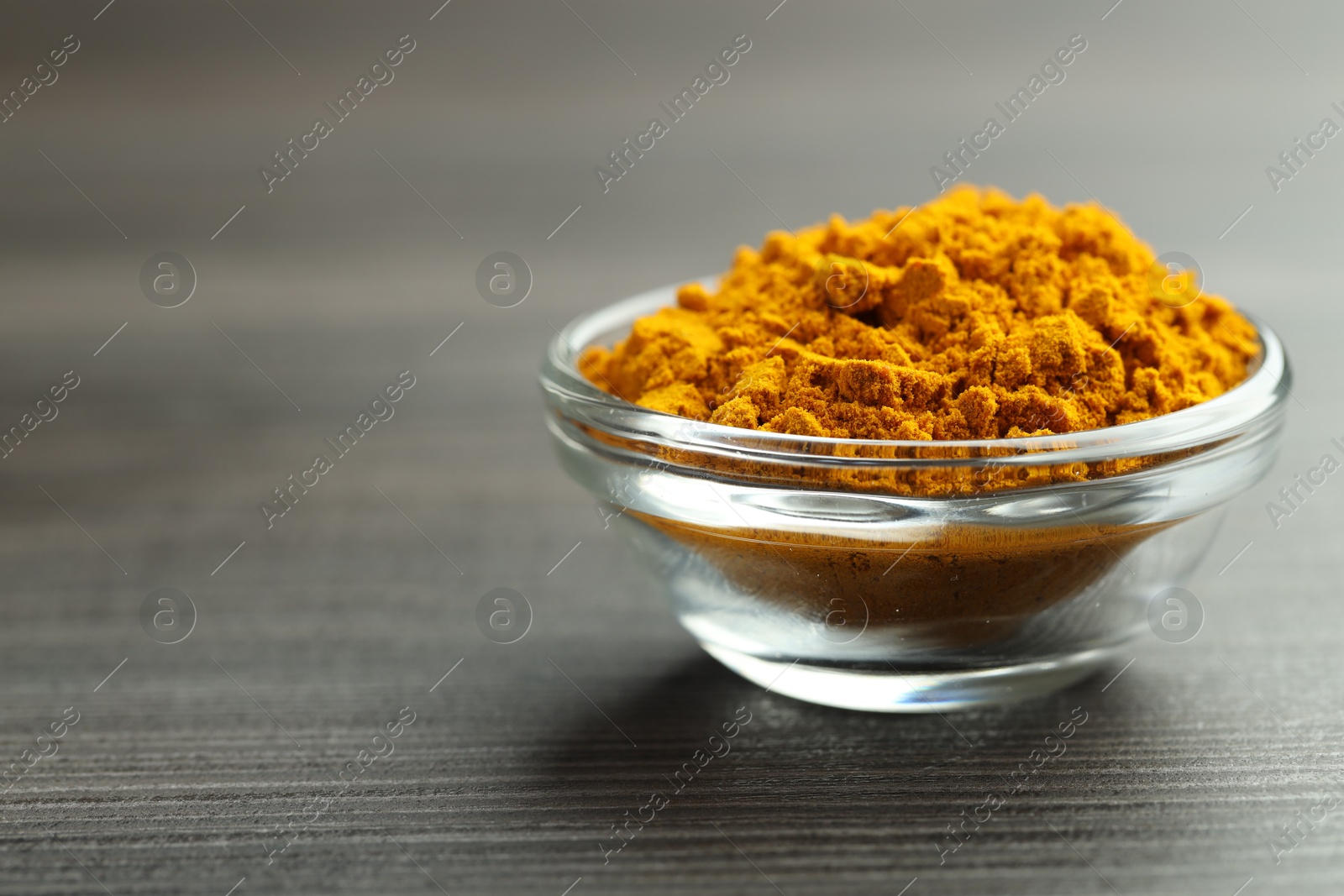 Photo of Turmeric powder in glass bowl on grey wooden table, closeup. Space for text