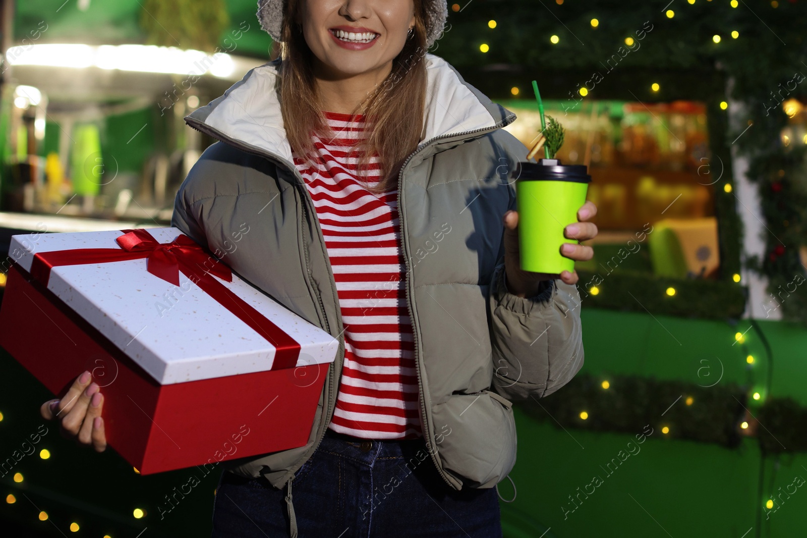 Photo of Happy woman with Christmas gift and paper cup near festive bus outdoors, closeup