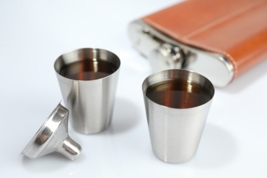 Photo of Hip flask, funnel and cups with whiskey on white table, closeup