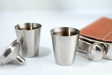 Photo of Hip flask, funnel and cups with whiskey on white table, closeup