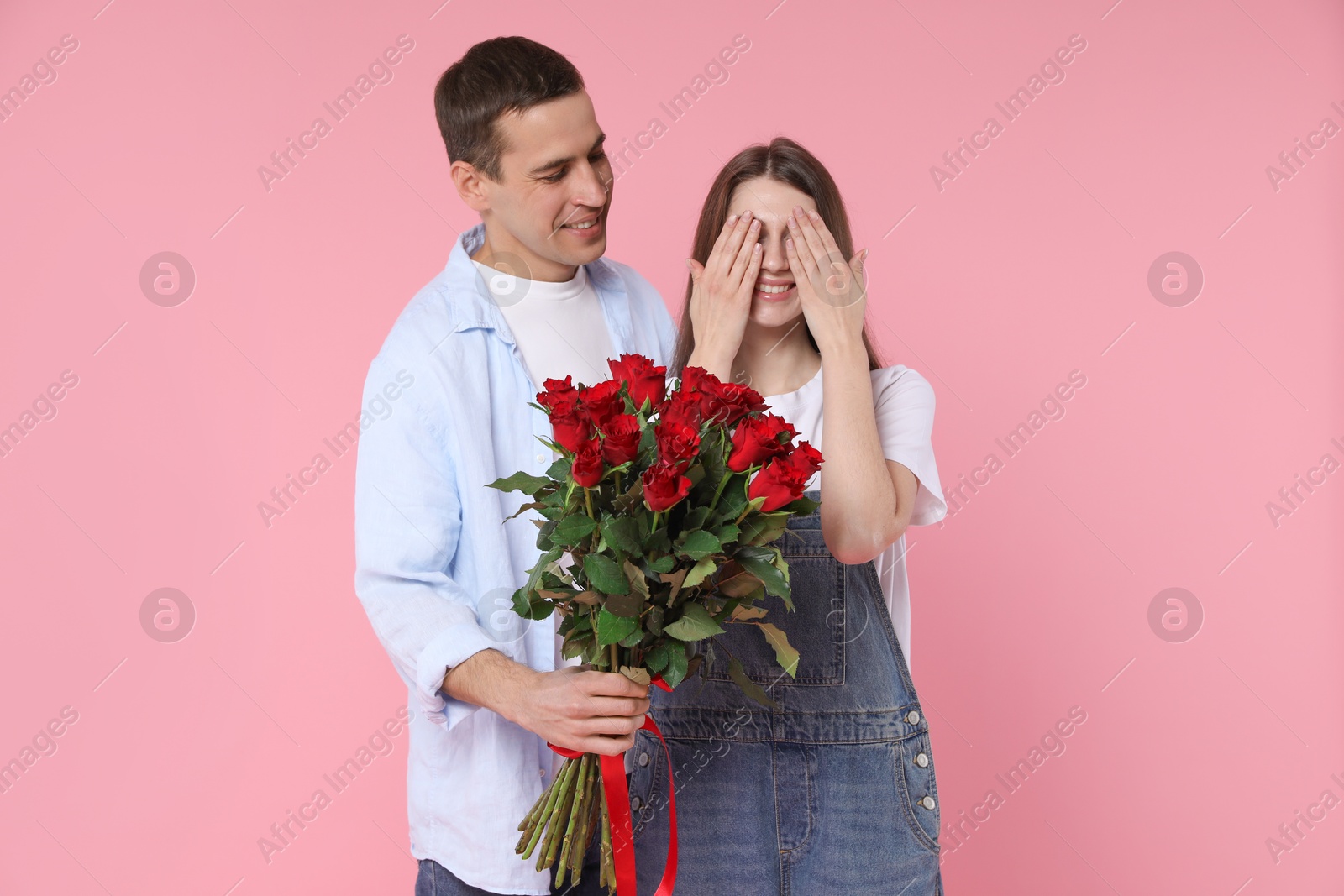 Photo of Lovely man surprising his girlfriend with bouquet of red roses on pink background. Valentine's day celebration