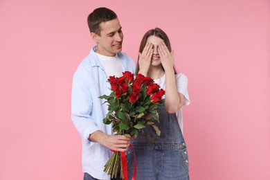 Photo of Lovely man surprising his girlfriend with bouquet of red roses on pink background. Valentine's day celebration