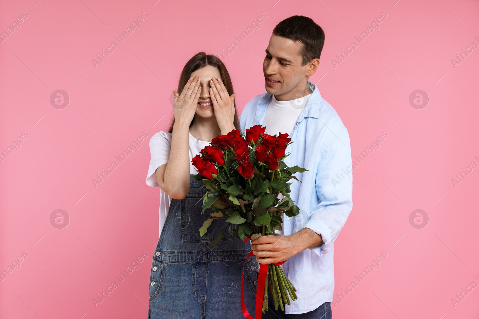 Photo of Lovely man surprising his girlfriend with bouquet of red roses on pink background. Valentine's day celebration