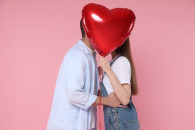 Photo of Lovely couple kissing behind heart shaped balloon on pink background. Valentine's day celebration