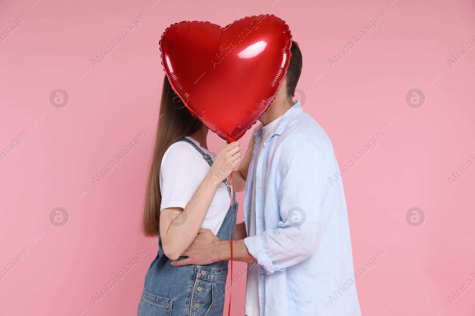 Photo of Lovely couple kissing behind heart shaped balloon on pink background. Valentine's day celebration