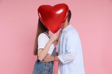 Photo of Lovely couple kissing behind heart shaped balloon on pink background. Valentine's day celebration