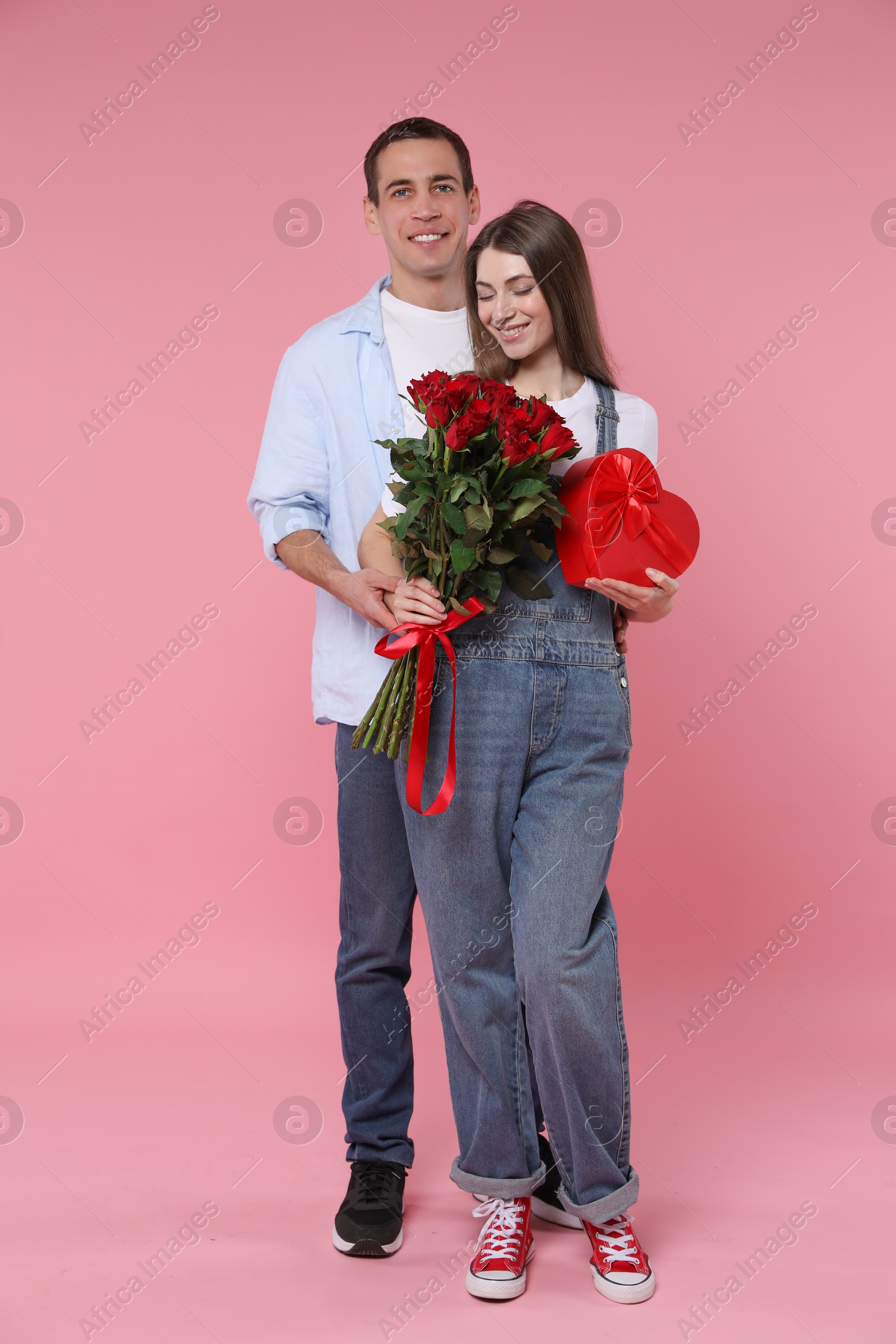 Photo of Lovely couple with bouquet of red roses and gift box on pink background. Valentine's day celebration