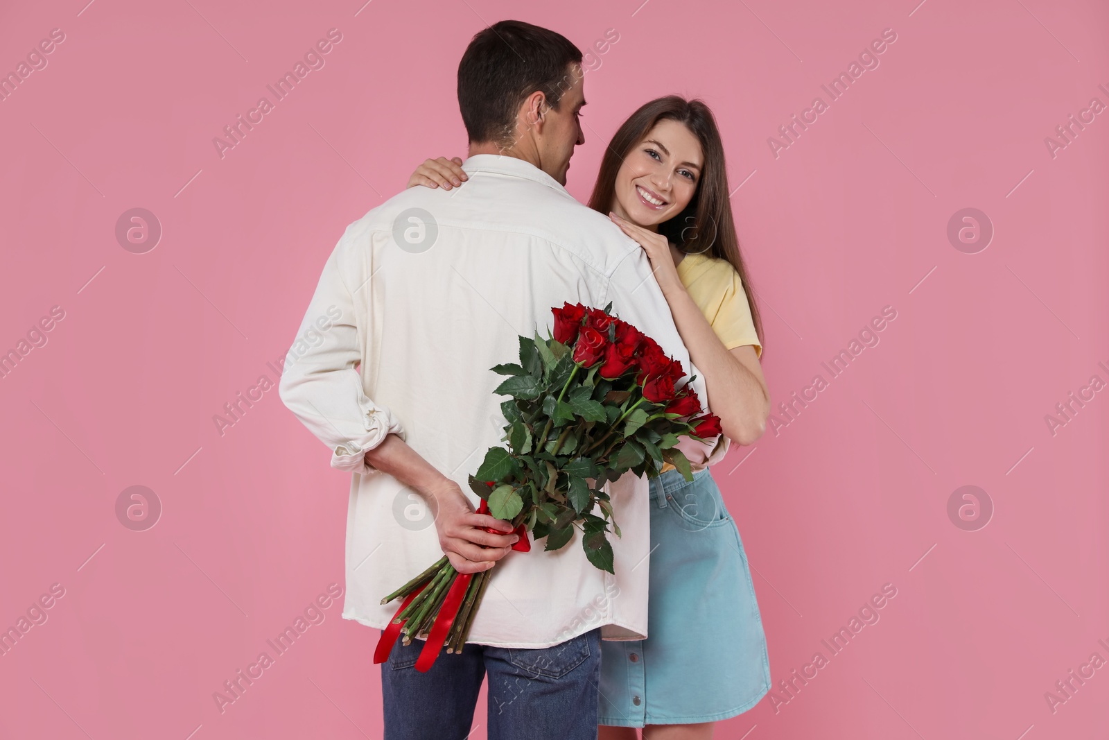 Photo of Lovely man hiding bouquet of red roses for his beloved woman on pink background. Valentine's day celebration