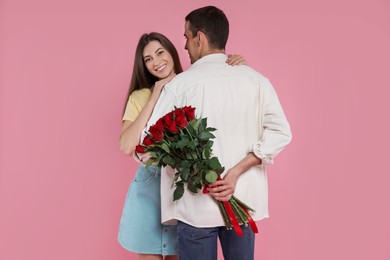 Photo of Lovely man hiding bouquet of red roses for his beloved woman on pink background. Valentine's day celebration