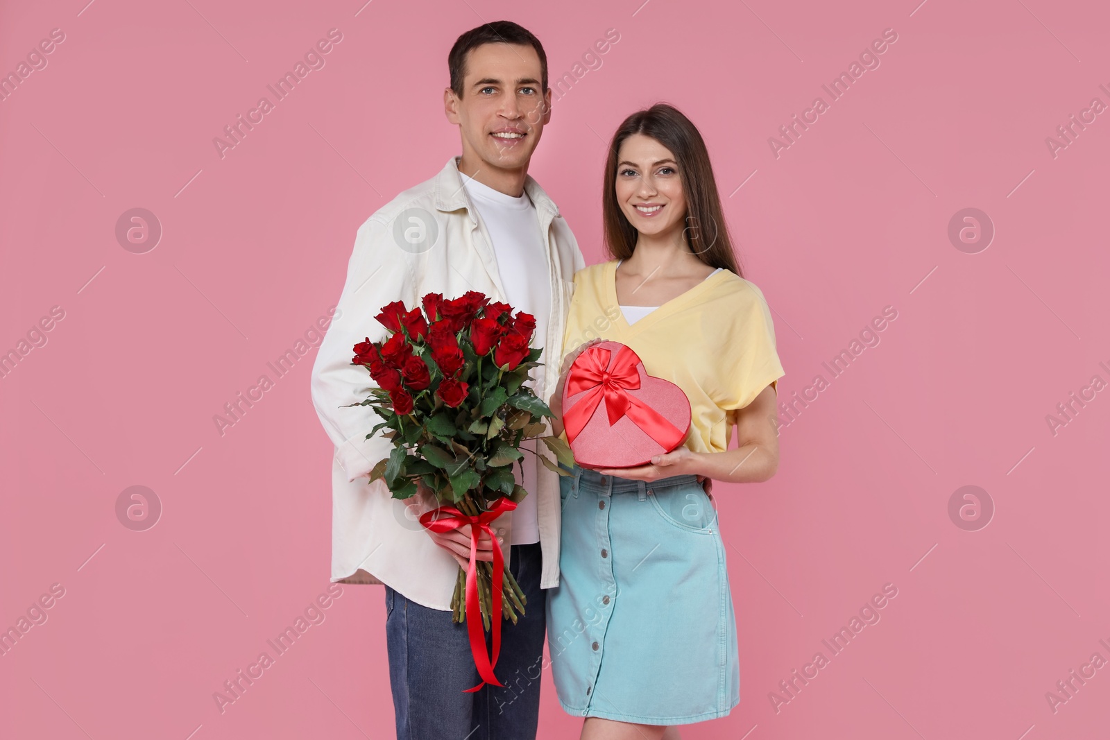 Photo of Lovely couple with bouquet of red roses and gift box on pink background. Valentine's day celebration