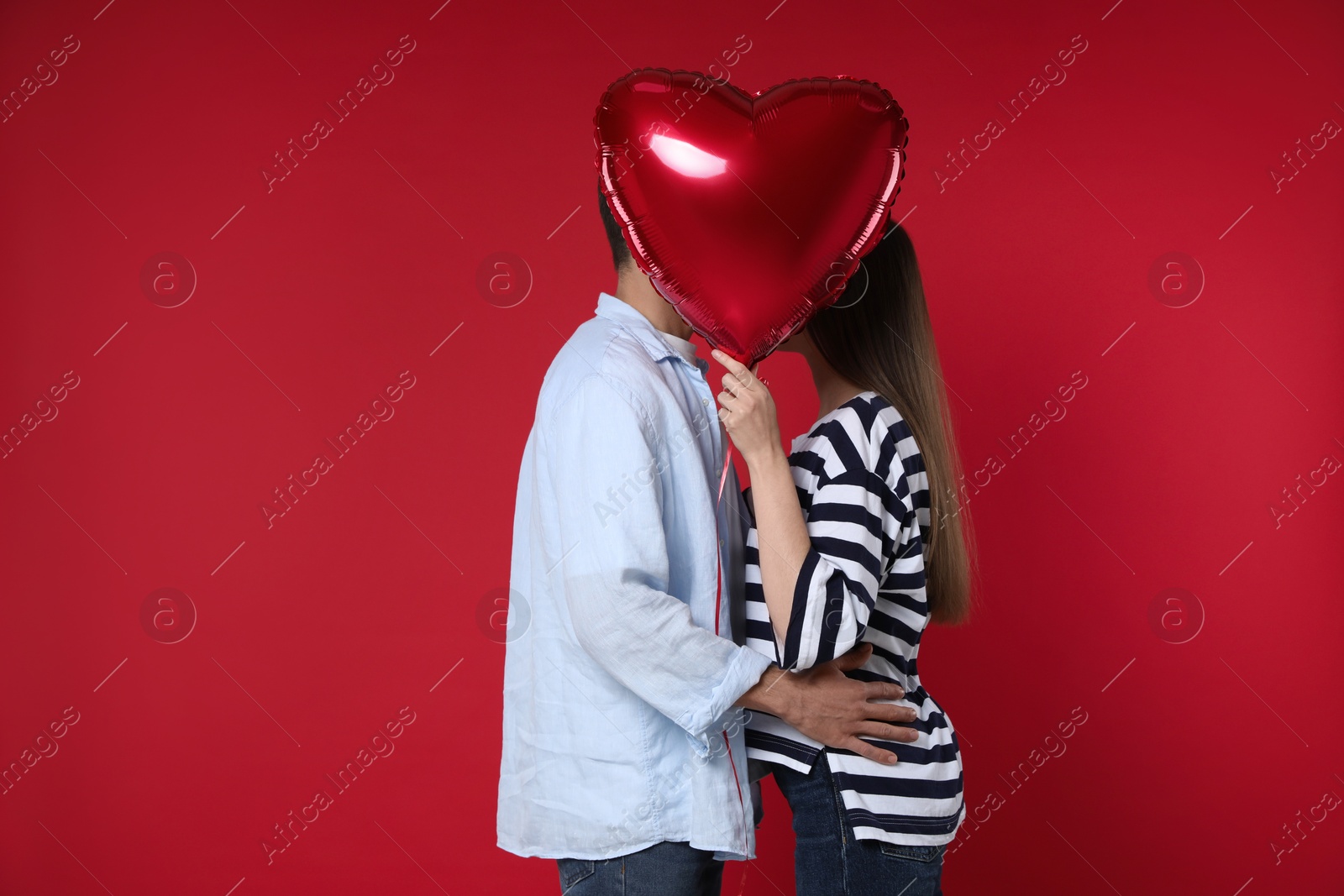 Photo of Lovely couple kissing behind heart shaped balloon on red background. Valentine's day celebration