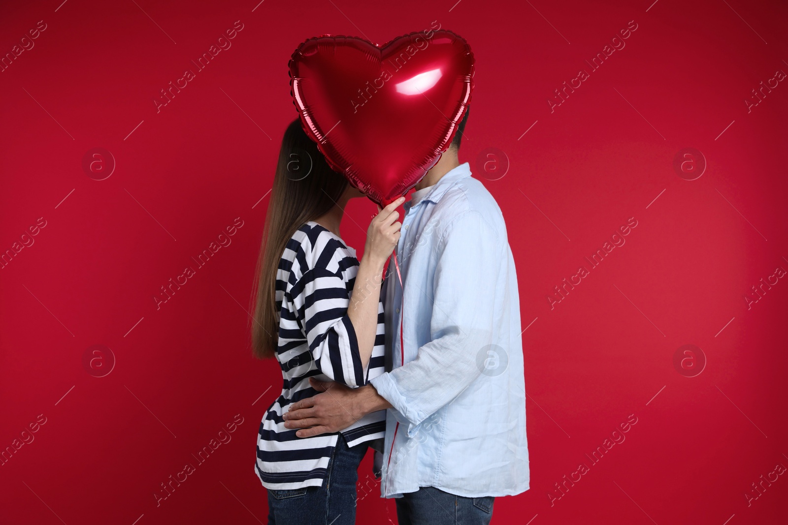 Photo of Lovely couple kissing behind heart shaped balloon on red background. Valentine's day celebration