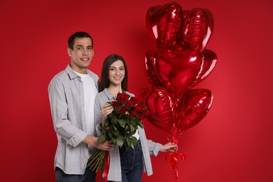 Photo of Lovely couple with heart shaped balloons and bouquet of roses on red background. Valentine's day celebration