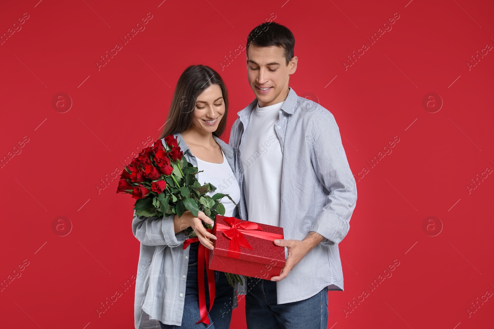 Photo of Lovely couple with bouquet of roses and gift box on red background. Valentine's day celebration