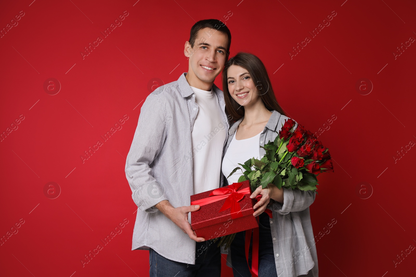 Photo of Lovely couple with bouquet of roses and gift box on red background. Valentine's day celebration