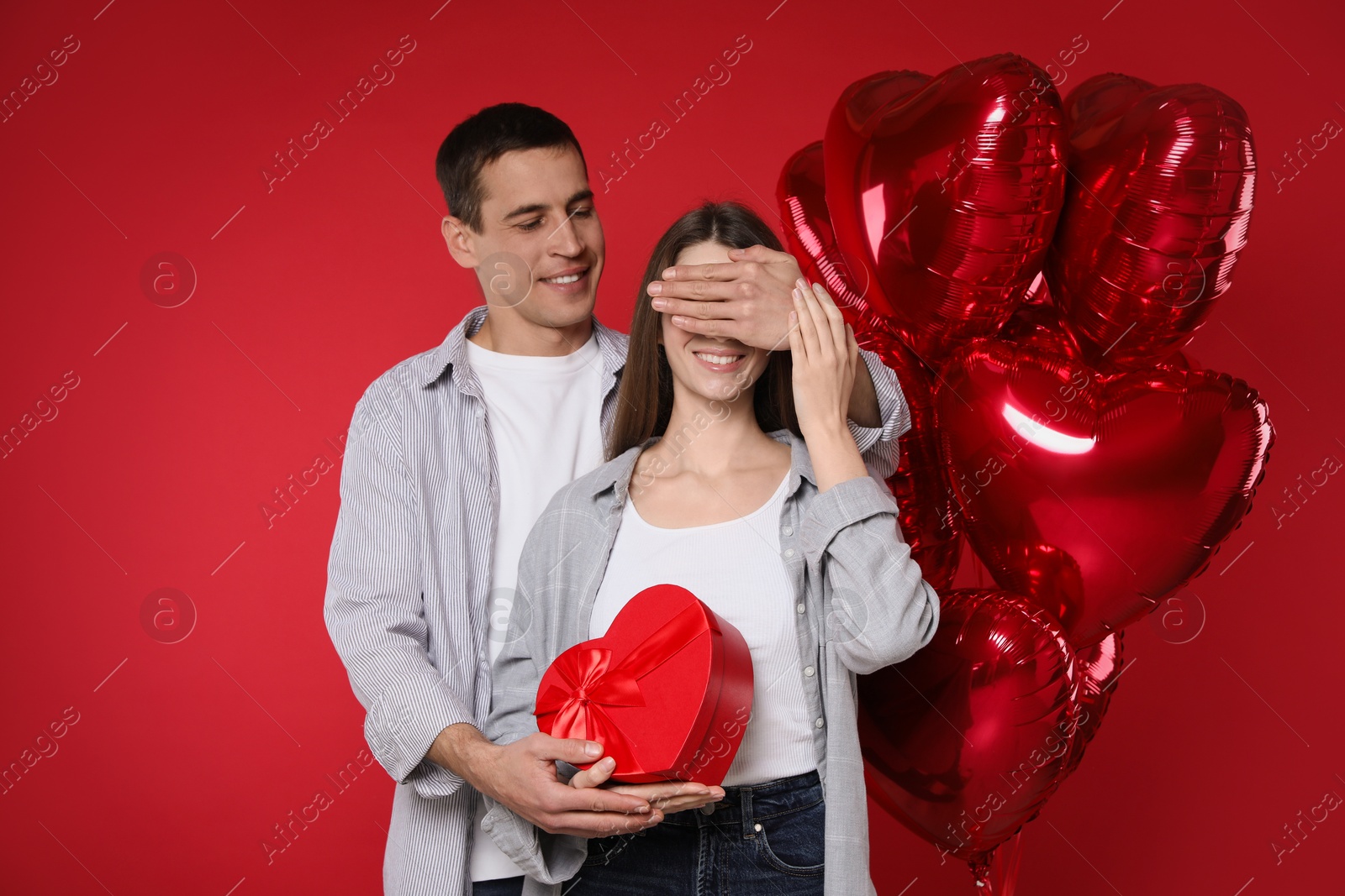 Photo of Lovely man surprising his girlfriend with Valentine's day gift near heart shaped balloons on red background