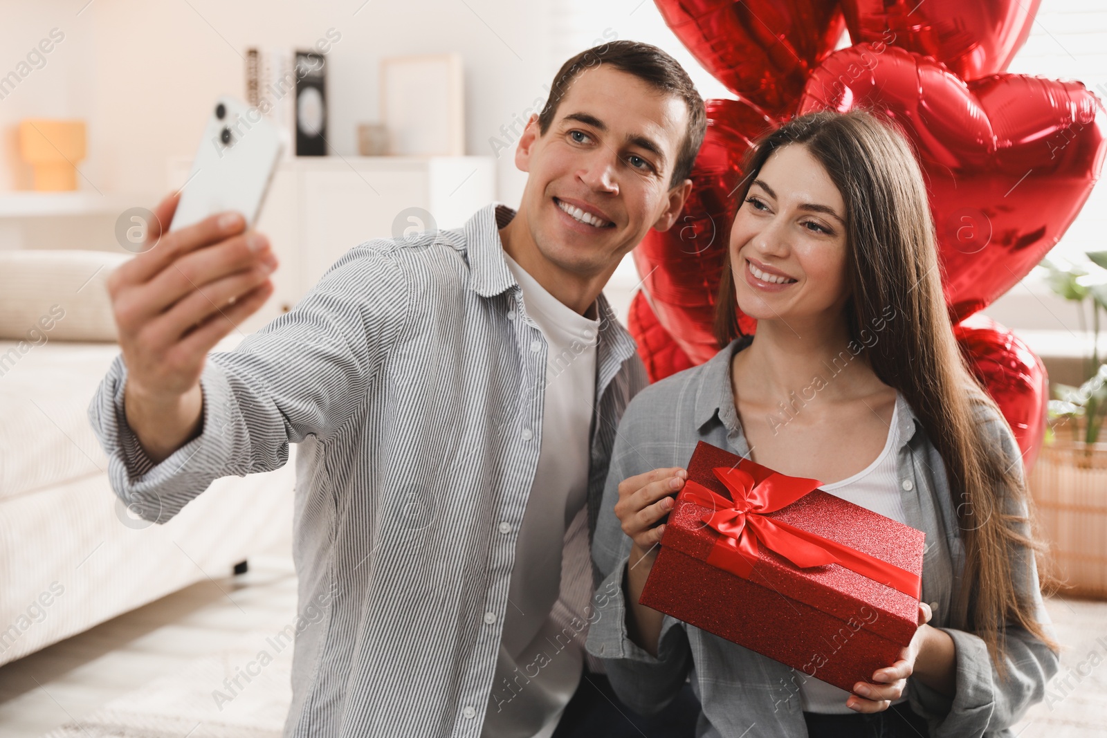 Photo of Lovely couple with gift box taking selfie near heart shaped balloons indoors. Valentine's day celebration