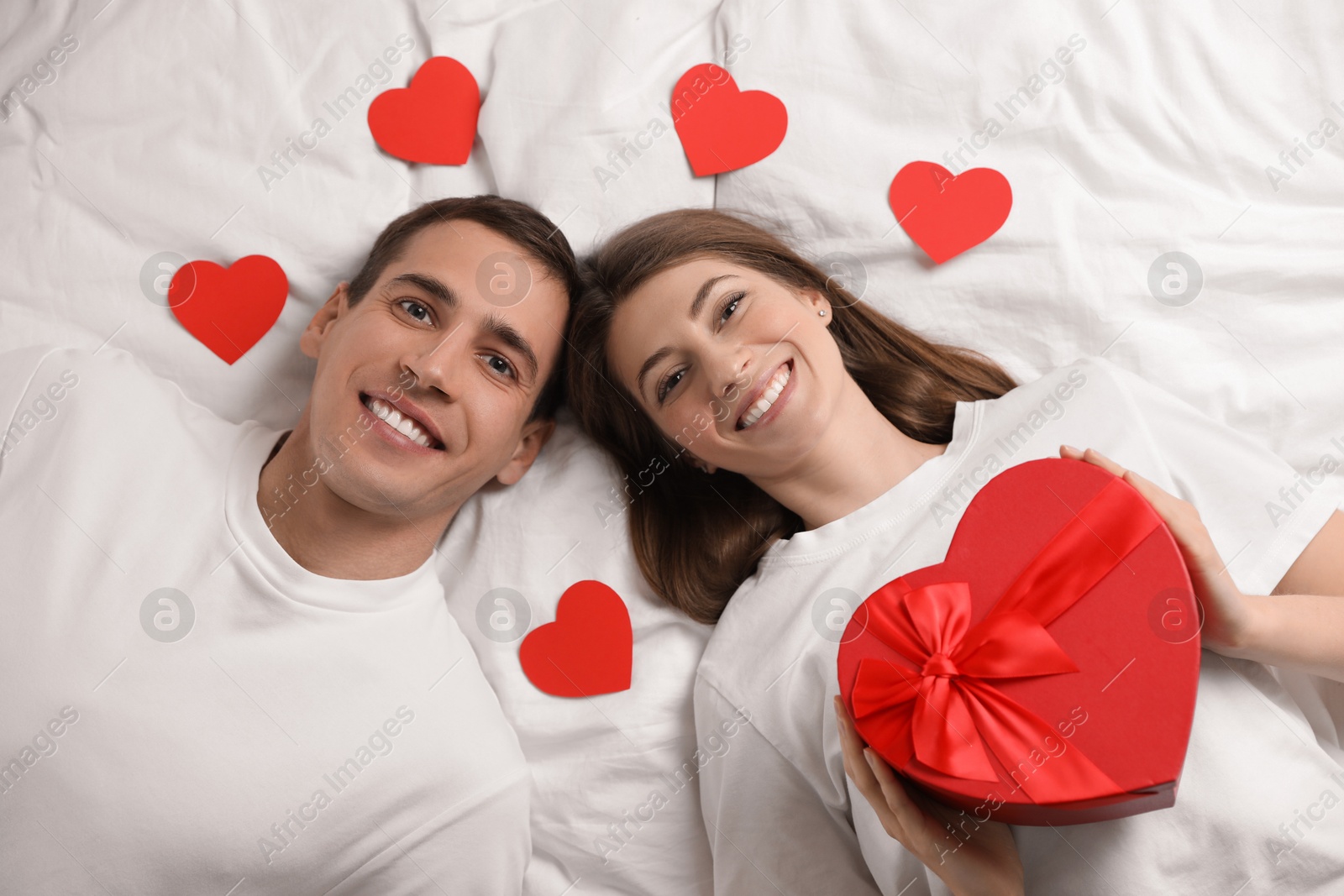 Photo of Lovely couple lying on bed with paper hearts and gift box, top view. Valentine's day celebration