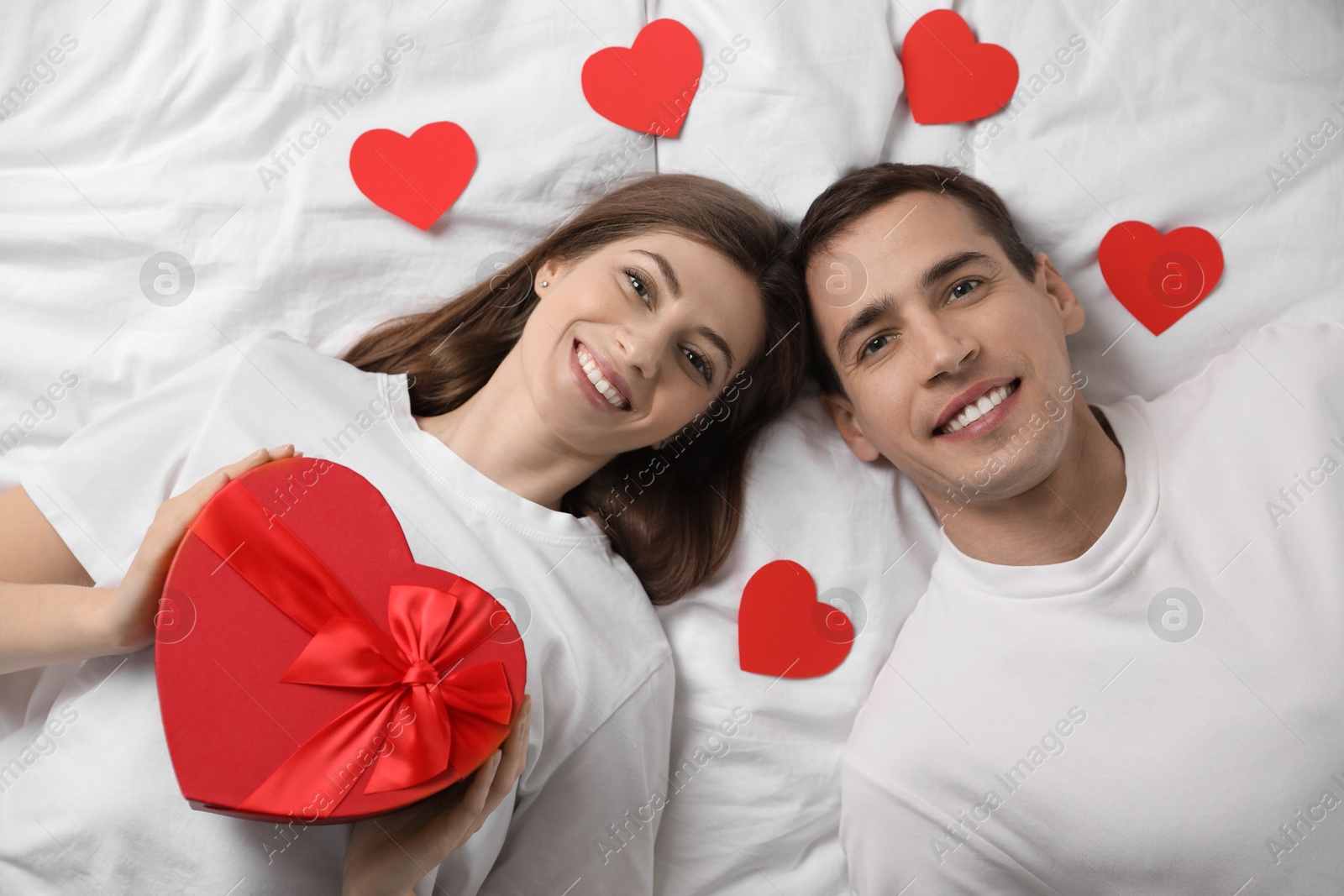 Photo of Lovely couple lying on bed with paper hearts and gift box, top view. Valentine's day celebration