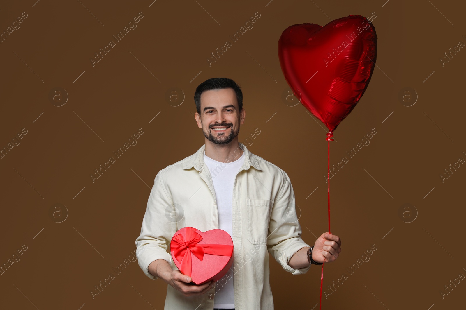 Photo of Happy Valentine's Day. Handsome man with heart shaped balloon and gift on brown background