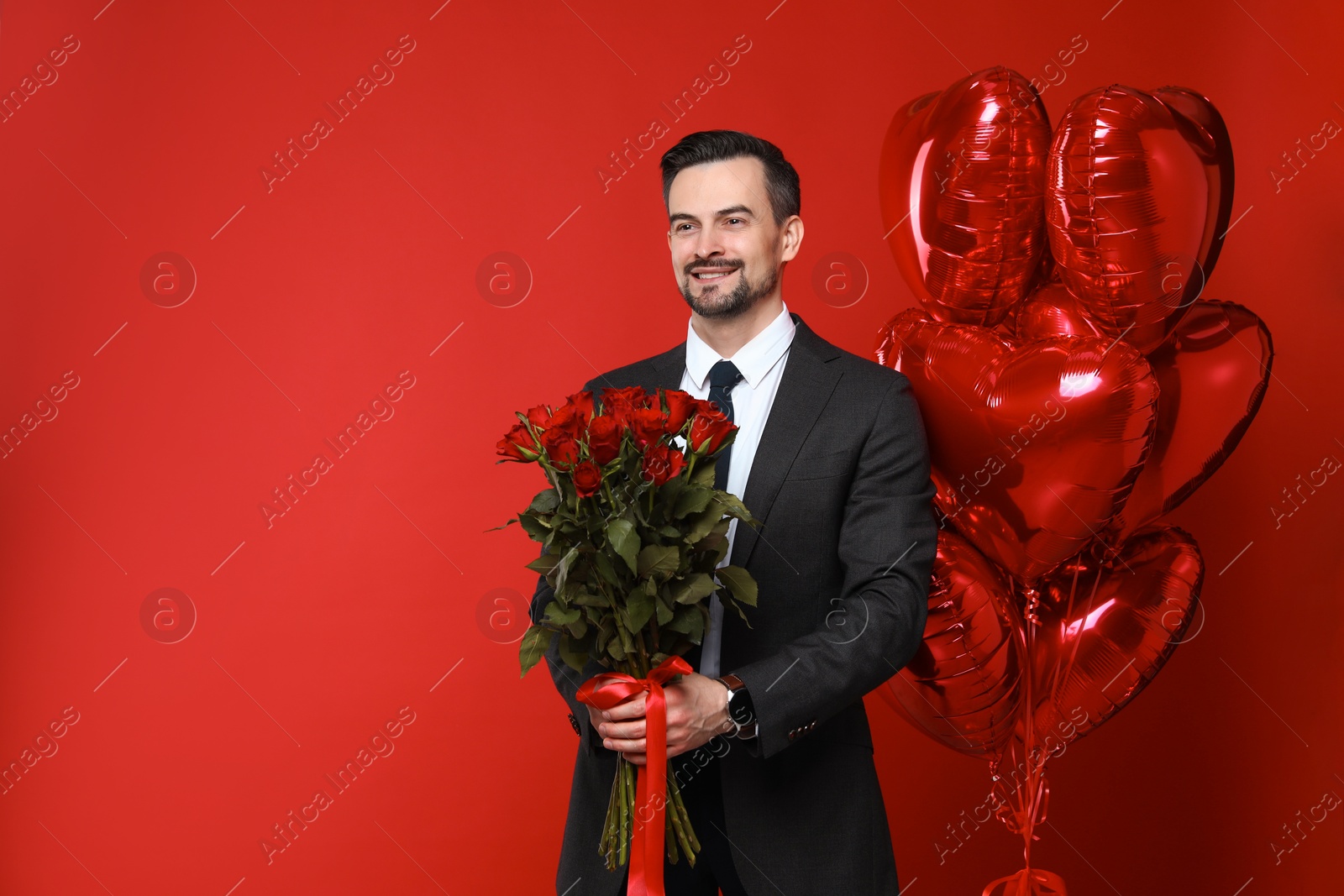 Photo of Happy Valentine's Day. Handsome man with heart shaped balloons and bouquet of roses on red background. Space for text