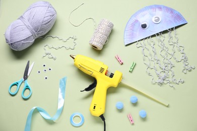 Photo of Hot glue gun and handicraft materials on pale olive background, flat lay