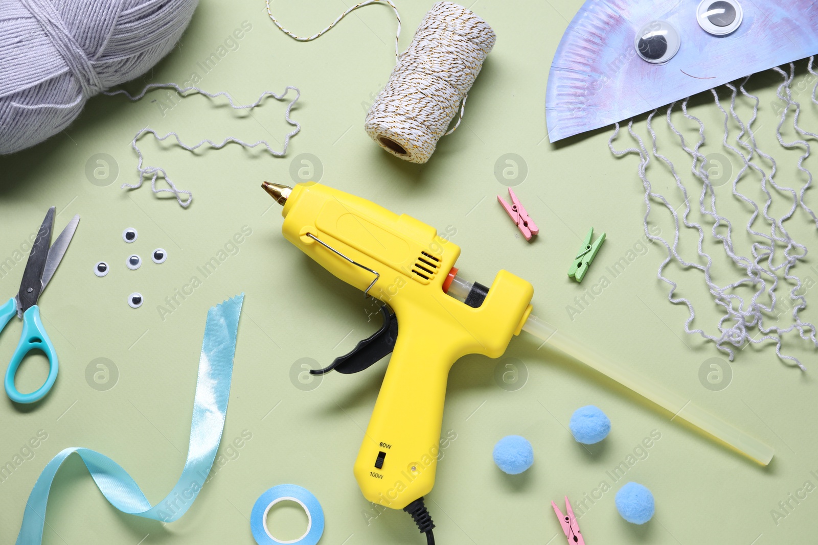 Photo of Hot glue gun and handicraft materials on pale olive background, flat lay