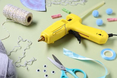 Photo of Hot glue gun and handicraft materials on pale olive background, closeup