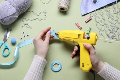 Photo of Woman with hot glue gun making craft on pale olive background, top view