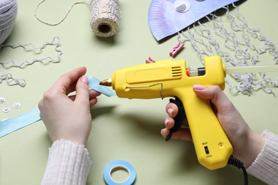 Photo of Woman with hot glue gun making craft on pale olive background, above view