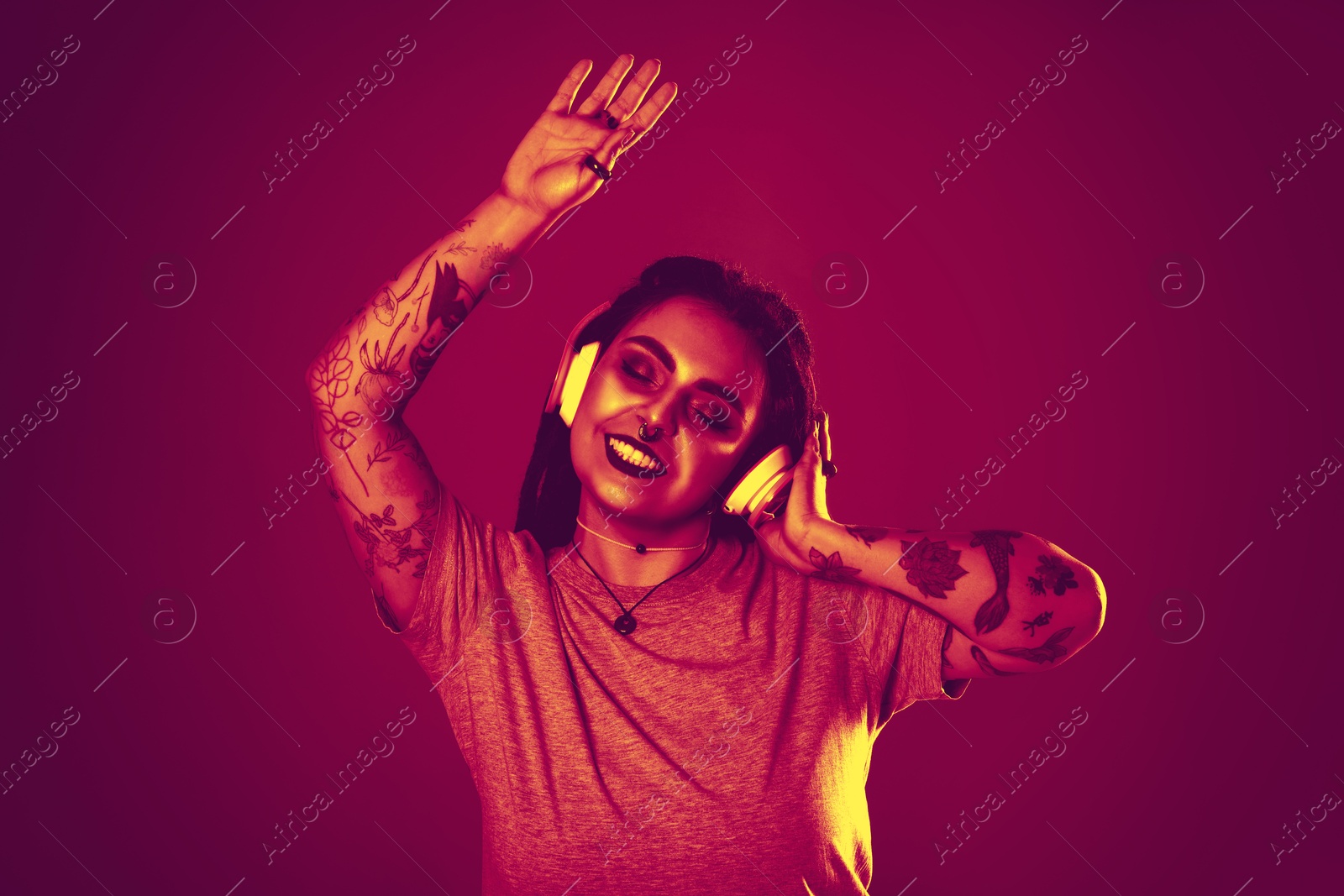 Image of Beautiful young woman with tattoos on arms, nose piercing and dreadlocks listening to music in neon light