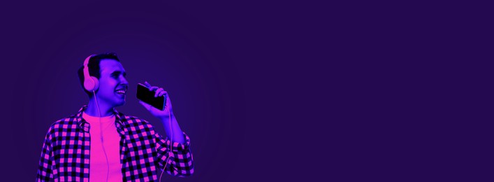 Image of Handsome young man with headphones and smartphone singing in neon light, space for text. Banner design