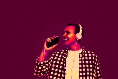 Image of Handsome young man with headphones and smartphone singing in neon light, space for text