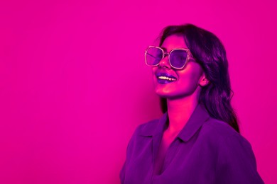 Image of Happy beautiful woman with stylish sunglasses in neon light, space for text